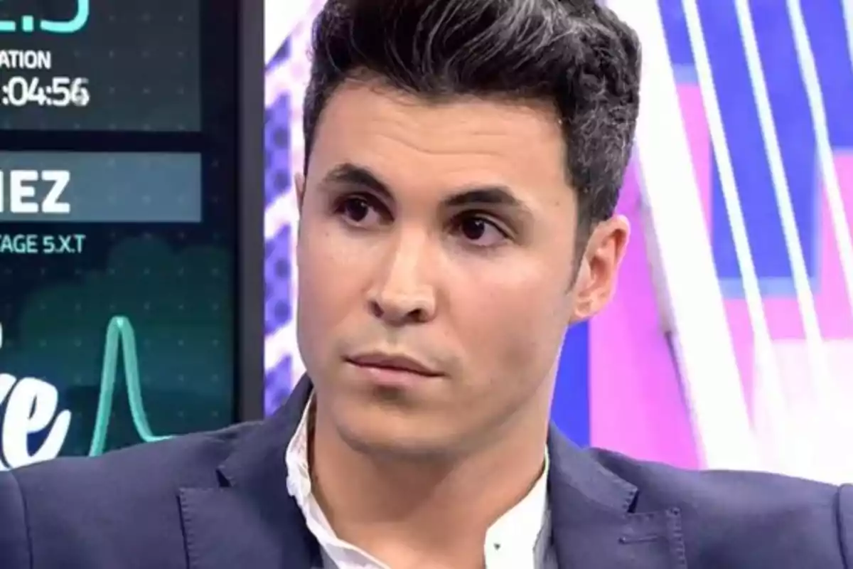 Hombre joven de cabello oscuro y corto con expresión seria, vistiendo saco azul y camisa blanca, en un set de televisión con luces y pantallas de fondo