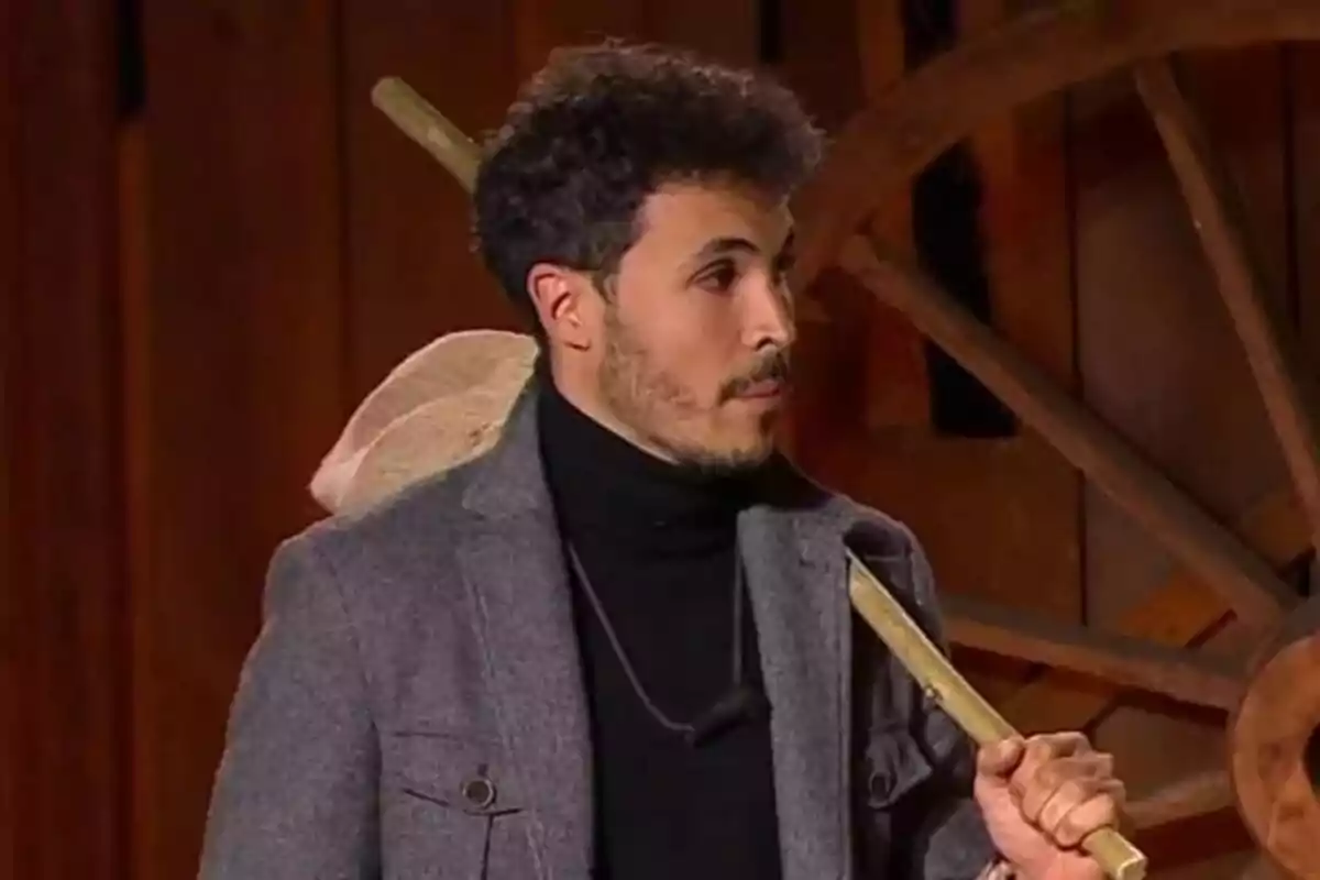 Hombre joven con barba y cabello rizado viste un abrigo gris y suéter negro mientras sostiene un palo de madera en un entorno rústico con una rueda de carreta al fondo