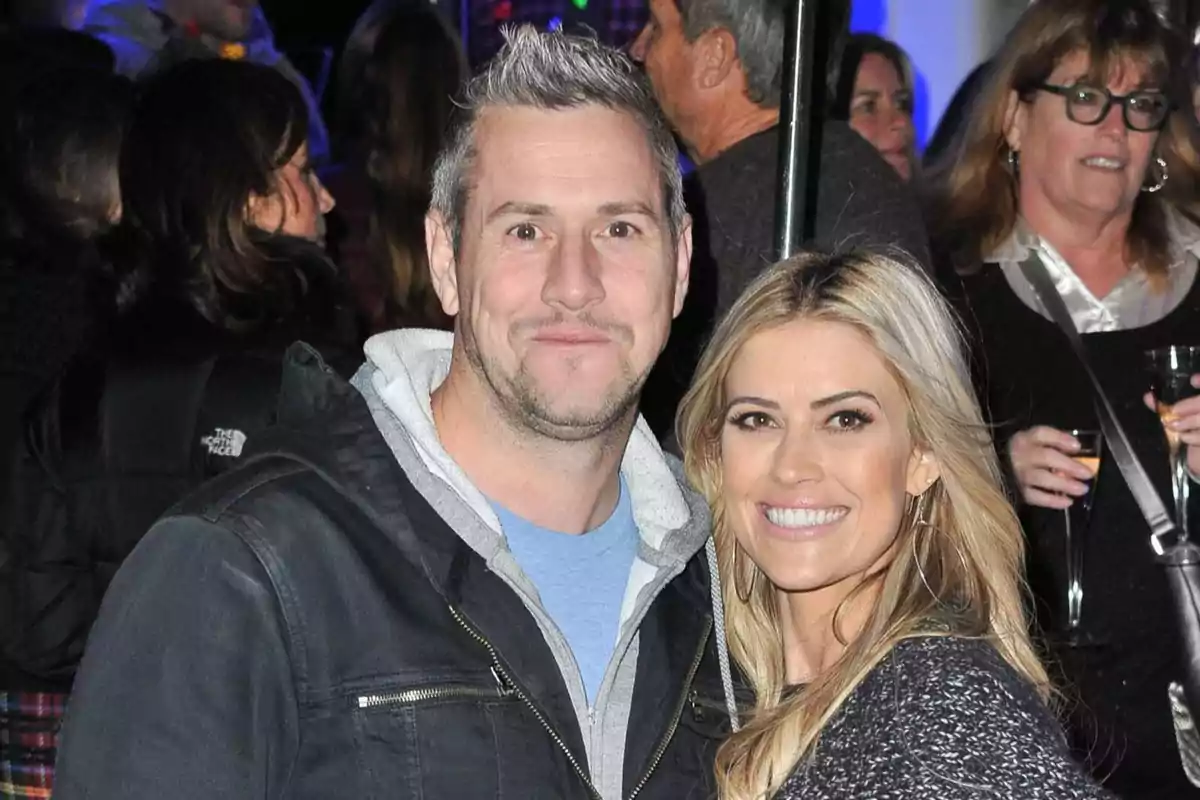 Una pareja sonríe mientras posa para una foto en un evento social con varias personas al fondo.