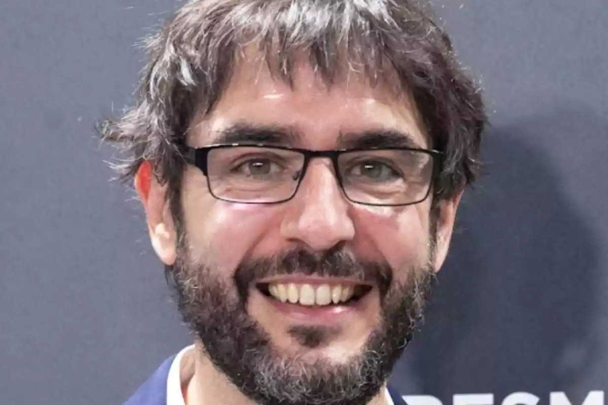 Hombre con barba y gafas sonriendo frente a un fondo gris