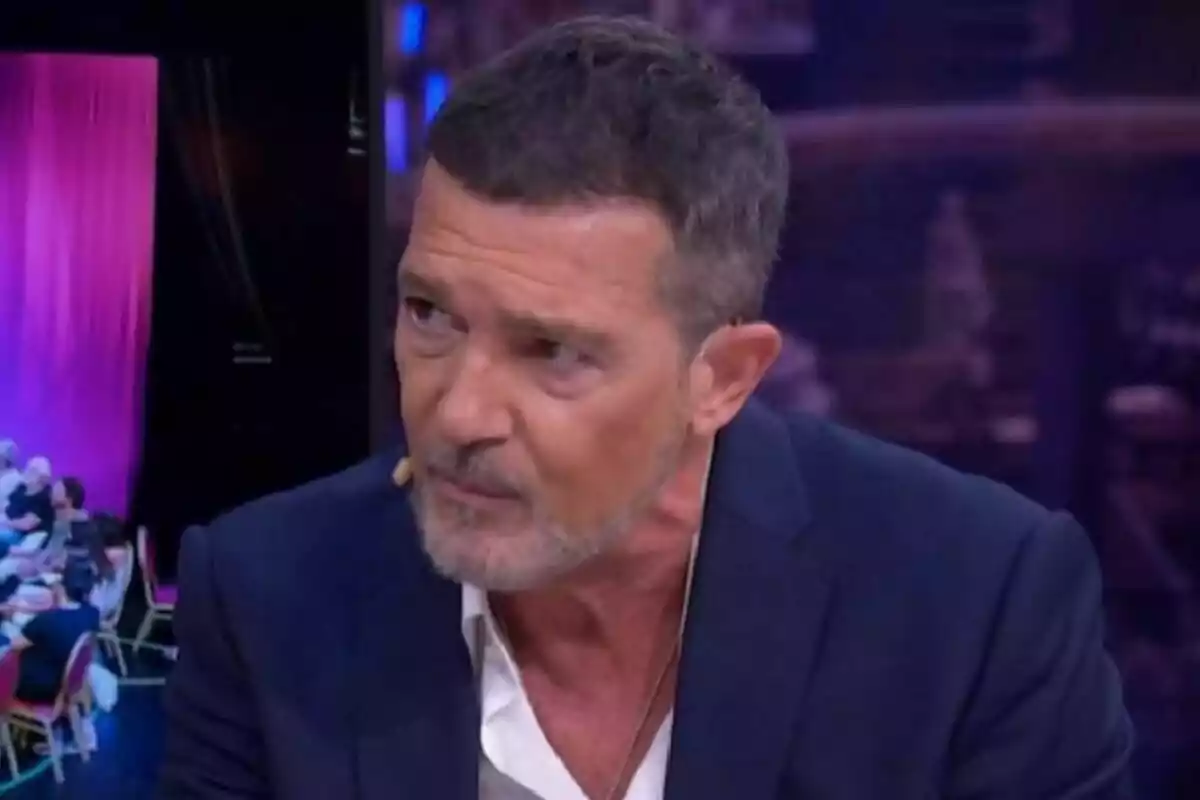 Antonio Banderas with a beard and short hair, dressed in a blue jacket and white shirt, speaking on El Hormiguero with a microphone in his ear. Antonio Banderas with a beard and short hair, dressed in a blue jacket and white shirt, speaking on El Hormiguero with a microphone in his ear.