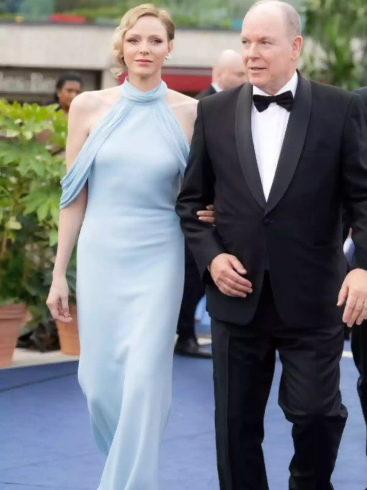 Una mujer con vestido azul claro y un hombre con esmoquin negro caminan juntos por una alfombra azul en un evento elegante al aire libre.