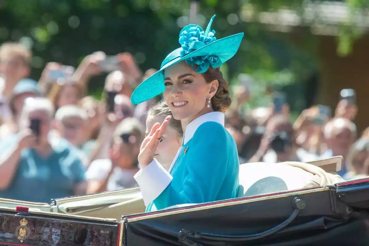 Mujer con sombrero azul sonríe y saluda desde un carruaje durante un evento al aire libre con público de fondo