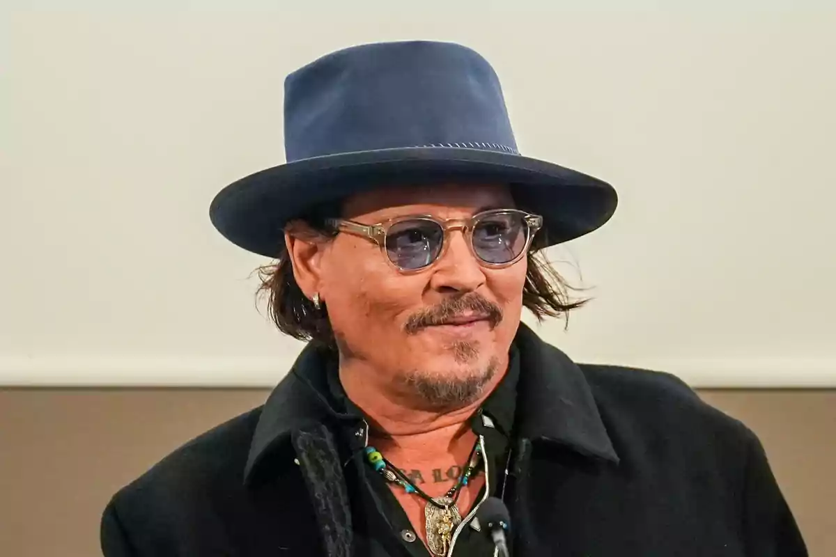 Johnny Depp with a hat and sunglasses smiles while wearing a dark coat.