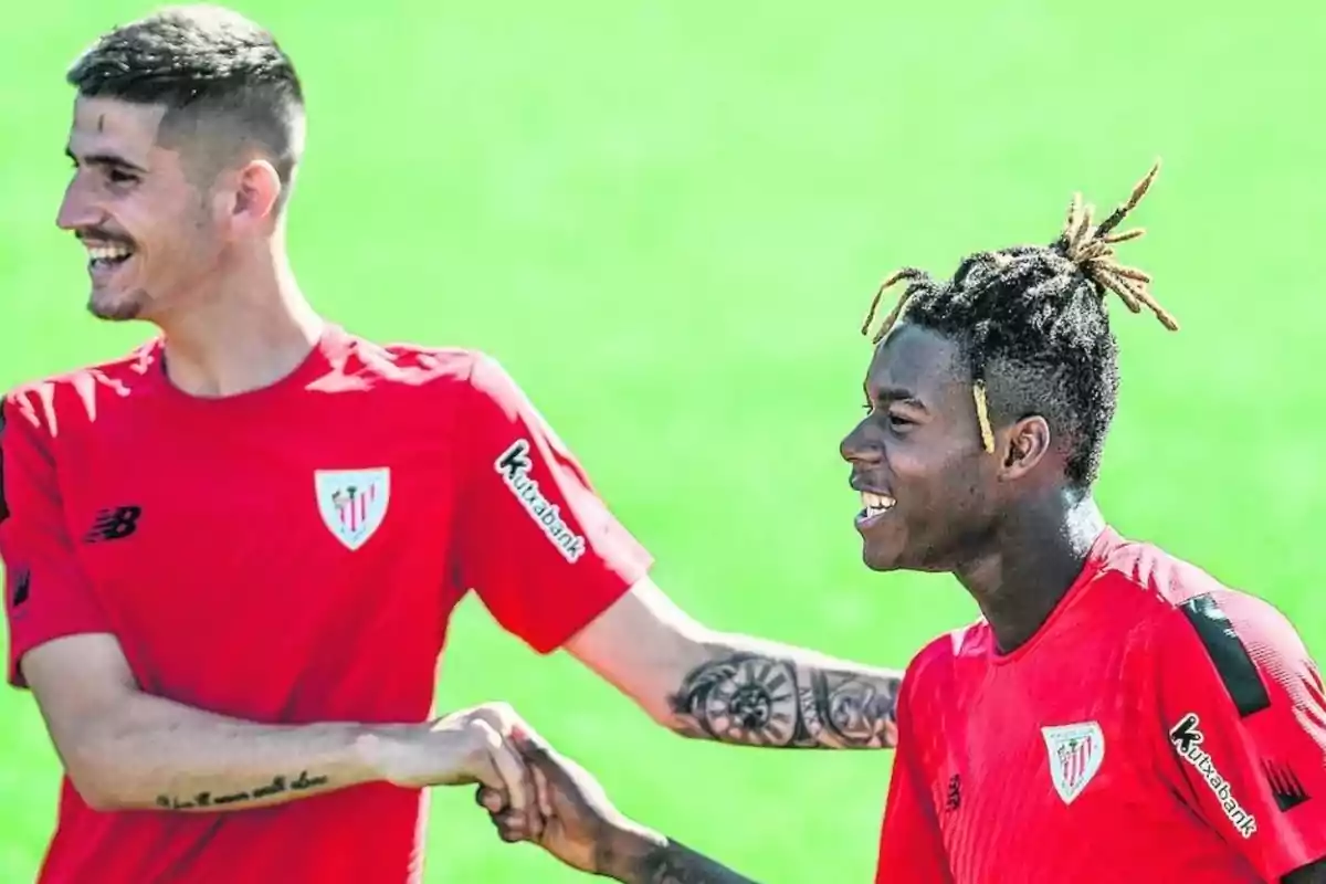 Dos jugadores de fútbol con camisetas rojas del Athletic Club se saludan sonrientes en un campo de entrenamiento.