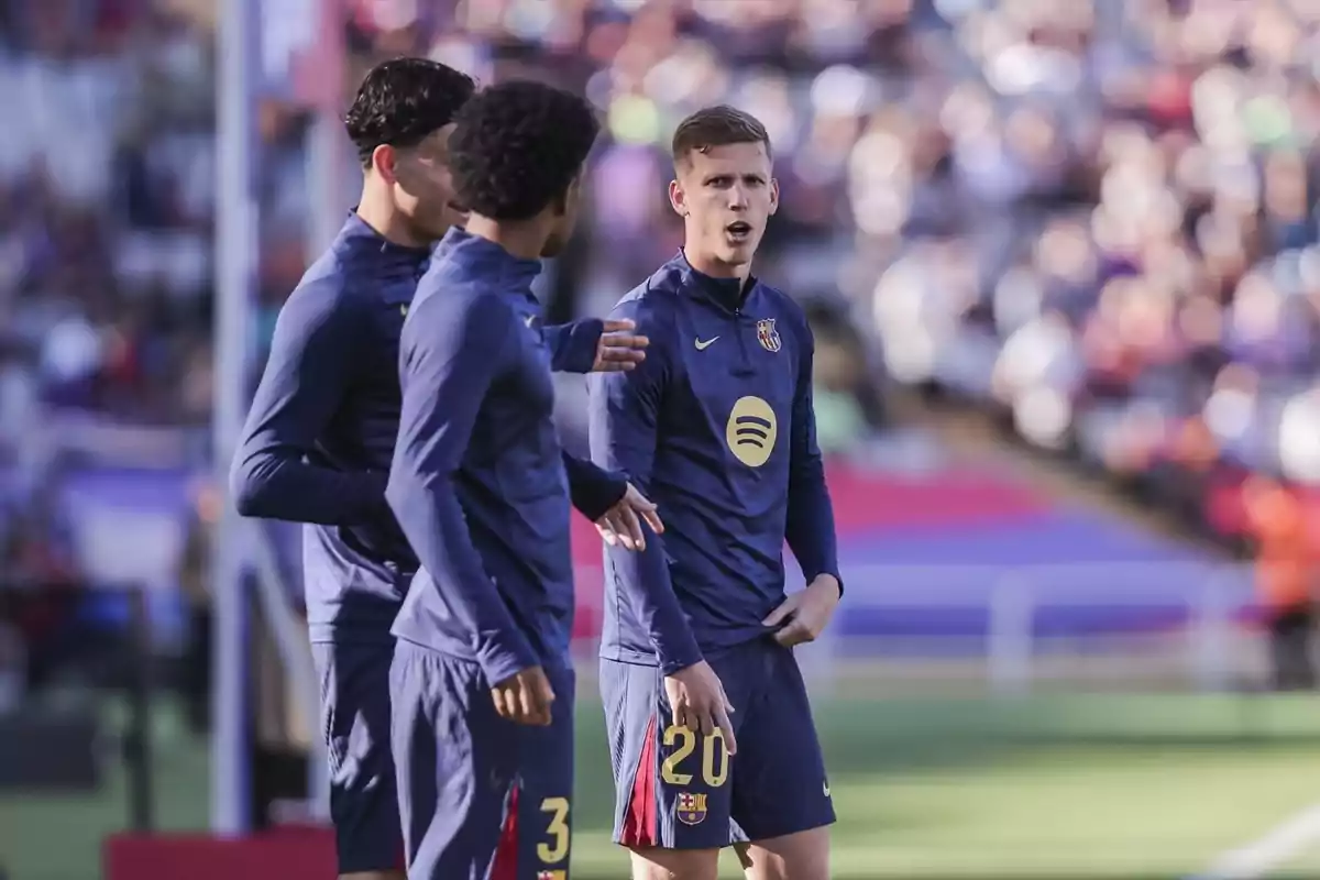 Tres jugadores de fútbol del FC Barcelona conversan en el campo durante un partido, vistiendo uniformes de entrenamiento azul oscuro.