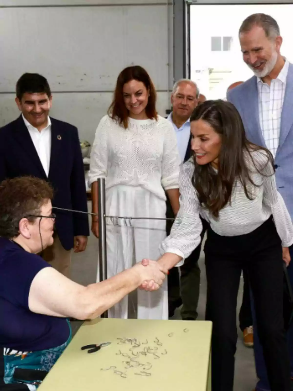 Una mujer sentada en una mesa saluda de mano a la reina Letizia de pie mientras varias personas observan sonrientes en un entorno interior