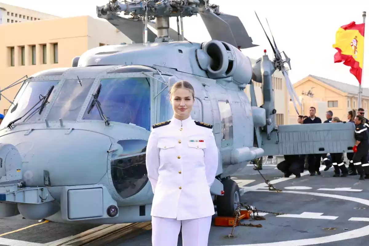Leonor con uniforme militar blanco de gala posando frente a un helicóptero en una base naval con bandera de España al fondo