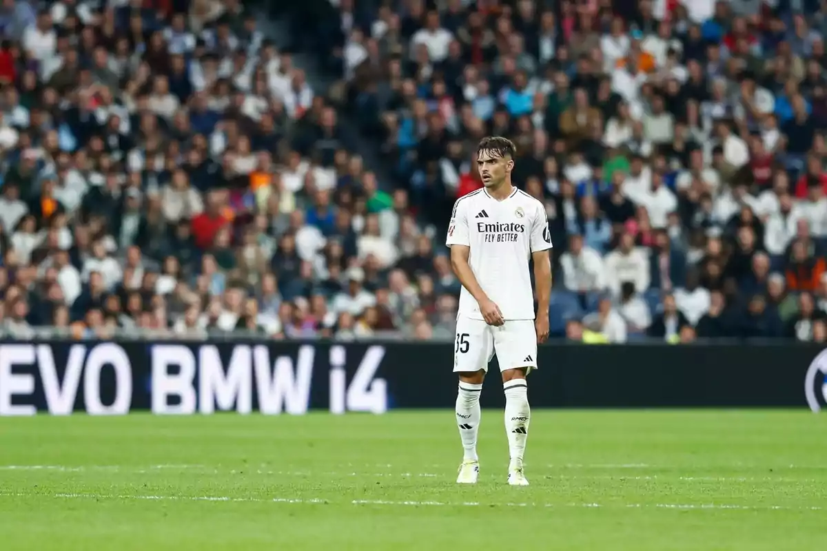 Un jugador de fútbol con el uniforme blanco del Real Madrid está de pie en el campo durante un partido, con una multitud de espectadores en el fondo. Un jugador de fútbol con el uniforme blanco del Real Madrid está de pie en el campo durante un partido, con una multitud de espectadores en el fondo.